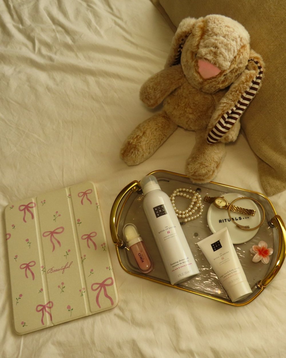 Flat lay com produtos Rituals numa bandeja dourada, peluche e capa de tablet com laços